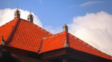 Geleneksel Bali çatılarının yakın plan görüntüsü. Canlı kırmızı fayanslar ve kabarık beyaz bulutlar ve mavi gökyüzü üzerine karmaşık taş oymalar..