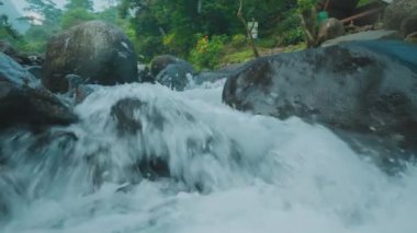 Pürüzsüz kayaların üzerinde akan bir nehrin yakın görüntüsü, berrak su çağlayarak akıyor ve su sıçrıyor. Bereketli yeşillik sahneyi çevreleyerek, sakin bir doğal ortam yaratıyor..