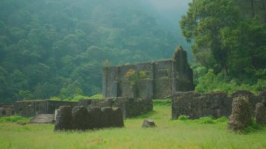 Arka planda sisli dağlar olan yemyeşil ormanlarla çevrili antik kalıntılar. Taş yapılar kısmen büyümüş, doğanın bu alandaki ıslahını gösteriyor..