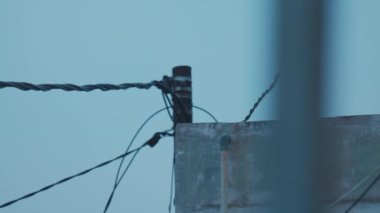 Mavi gökyüzüne karşı elektrik kabloları ve direği olan bir çatı manzarası. Görüntü binanın dokusunu ve kabloların düzenini yakalıyor ve endüstriyel bir estetik yaratıyor..