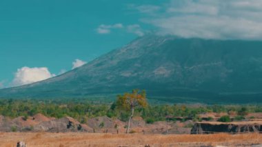 Bereketli yeşillik ve kuru bir manzarayla çevrili büyük bir volkanın manzarası. Gökyüzü birkaç bulutla berrak, görkemli dağı aydınlatıyor..