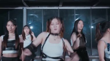 Bir grup dansçı modern bir stüdyoda koreografi gösterisi yapıyor. Beyaz bluz ve siyah aksesuarlar giyen merkez dansçı, diğerleri arka planda hareket ederken dinamik bir poz verir.. 