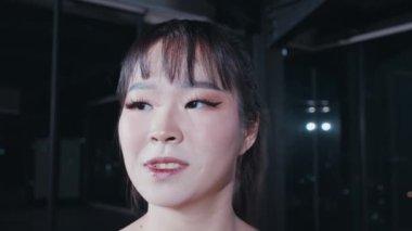 Uzun siyah saçlı ve belirgin makyajlı, kanatlı göz kalemi olan gülümseyen genç bir kadının yakın plan portresi. Arka plan hafifçe aydınlatılmış, sıcak bir atmosfer yaratıyor..