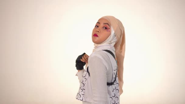 Une jeune femme élégante portant un hijab et une tenue à la mode, posant en toute confiance avec une expression ludique. Son maquillage est vibrant, avec un maquillage coloré pour les yeux et un rouge à lèvres audacieux. Le fond est clair, mettant l'accent sur sa tenue et posant.