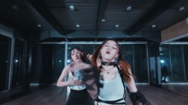 Geniş pencereli modern bir stüdyoda enerjik bir şekilde dans eden bir grup genç kadın. Şık kıyafetler giyiyorlar, çeşitli dans figürleri sergiliyorlar. Atmosfer hareketli ve dinamik, hareket ve ifade odaklı..