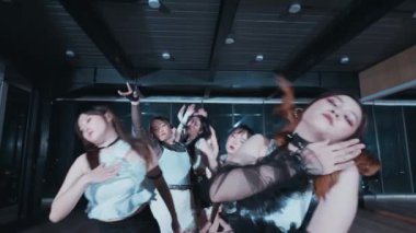 Bir grup dansçı stüdyoda gösteri yapıyor, kolları havada dinamik pozlar veriyorlar. Dansçılar şık kıyafetler giyip enerjik bir atmosfer yaratıyorlar. Arka planda geniş pencereli modern bir dans stüdyosu yer alıyor..