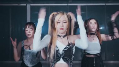 Bir grup dansçı modern bir stüdyoda koreografi gösterisi yapıyor. Beyaz bluz ve siyah aksesuarlar giyen merkez dansçı, diğerleri arka planda hareket ederken dinamik bir poz verir.. 