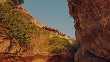 Aşağıdan çekilen verimli yeşilliklerle çevrili uçurum kenarındaki evlerin manzarası. Günbatımının ılık ışığı kayaların ve yaprakların üzerinde altın bir renk bırakır ve sakin bir atmosfer yaratır..