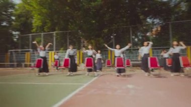 Bir grup üniformalı insan, açık hava basketbol sahasında yarı daire şeklinde dizilmiş sandalyelerde oturuyor. Arka planda ağaçlar ve çitler var..