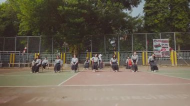 Geleneksel giysiler içinde bir grup dansçı bir basketbol sahasında koreografili bir gösteri yapıyorlar. Sandalyelerle sıralanmış, senkronize hareketleri sergiliyorlar.. 