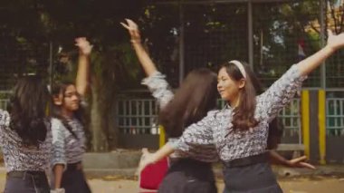 Bir grup kız açık havada dans ediyor, bazıları hareket halinde ve diğerleri oturuyor. Kurulumda sandalyeler ve renkli afişlerle bir zemin yer alıyor. Kızlar uyumlu kıyafetler giyiyorlar, canlı ve enerjik bir atmosfer sergiliyorlar..