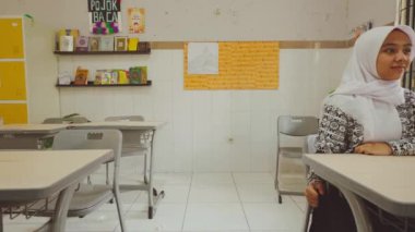 Sıralarda oturan öğrencilerle dolu bir sınıf, ön tarafa bakıyor. Öğrenciler özenli ve meşgul görünüyorlar. Sınıfın duvarlarında sarı dolaplar ve posterler var..