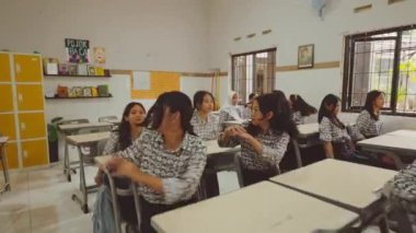 Aynı üniformayı giyen, sohbet ve aktivitelerle meşgul öğrencilerle dolu bir sınıf. Odanın duvarlarında masalar, sandalyeler ve eğitim posterleri var..