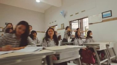 Üniformalı öğrencilerin olduğu bir sınıf sahnesi, bazıları ayakta ve bazıları sıralarda oturuyor. Odanın beyaz duvarları, parmaklıklı pencereleri ve duvarında bir vantilatör var..