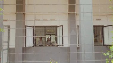 Bir grup insan pencerenin parmaklıklarından dışarı uzanıyor, neşeli ya da heyecanlı bir şekilde görünüyor. Ayarlar parmaklıklı pencereli bir binaya benziyor..