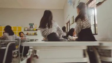 Sınıfta bir grup öğrenci sıralarında oturuyor ve birbirleriyle etkileşime geçiyorlar. Sınıfta sarı dolaplar ve arka planda bir kitaplık var..