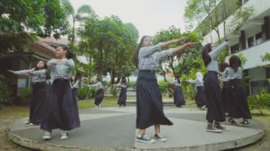 Bir grup üniformalı öğrenci, okul bahçesinde ağaçlar ve binalarla çevrili bir şekilde dans ediyorlar..