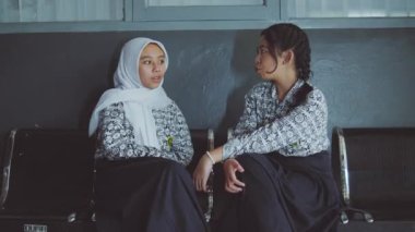 Aynı desenli gömlekleri ve uzun etekleri olan iki kadın metal sandalyelere oturup sohbet ediyorlar. Bir kadın tesettür takıyor. Başka bir kişi kısmen görünür, görüntünün sol tarafında hızla hareket ediyor..