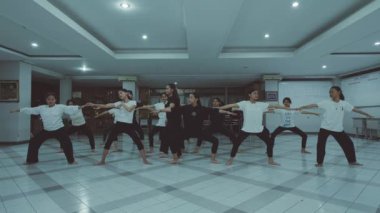 Kapalı bir odada fayanslı döşemeleri olan bir grup insan yoga yapıyor. Kollarını açıp denge ve duruşlarına odaklanarak geniş bir duruş sergiliyorlar..