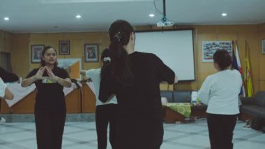 Tahta duvarları ve projektör ekranı olan bir odada dans veya egzersiz sınıfına katılan bir grup kadın..