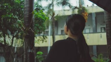 Uzun saçlı, at kuyruklu bir insan yağmurda dışarıda kameradan uzak duruyor. Arka planda bir bina ve ağaçlar görünüyor..