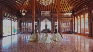 Geleneksel kıyafetli üç dansçı güzel dekore edilmiş ahşap bir salonda karmaşık oymalar ve bir avize ile gösteri yapıyorlar..