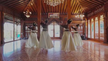 Bir grup dansçı, güzel dekore edilmiş ahşap bir salonda geleneksel giysiler içinde karmaşık oymalar ve bir avize ile gösteri yapıyorlar..