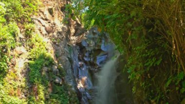 Bereketli yeşilliklerle çevrili kayalık bir uçurumdan şelalenin şelalesi düşüyor. Güneş ışığı ağaçların arasından süzülüp sakin ve sakin bir atmosfer yaratır..