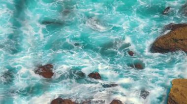 Beyaz köpüklü dalgaların kayalık kıyılara çarptığı turkuaz okyanus sularının havadan görüntüsü. Canlı renkler dinamik ve dinamik bir doğal sahne yaratır..