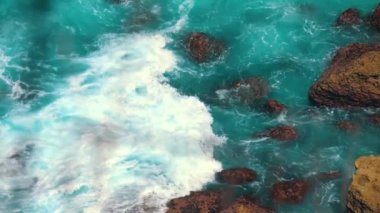 Turkuaz okyanus dalgalarının hava manzarası kayalık kıyılara çarpıyor, beyaz köpük ve su sıçratıyor. Suyun canlı renkleri engebeli kayalarla çok güzel tezat oluşturuyor..