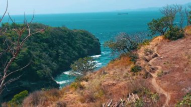 Kayalık bir kıyı şeridi, yemyeşil yemyeşil ve berrak mavi bir okyanusa sahip manzaralı bir sahil manzarası. Manzara, ufukta uzak tepeler ve suda seyreden bir gemiyle, uçurum kenarında dolambaçlı bir yol içerir..