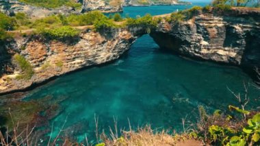Berrak turkuaz suların üzerinde doğal bir kaya kemerinin bulunduğu, yemyeşil ve kayalık uçurumlarla çevrili çarpıcı bir kıyı manzarası. Sahne doğanın güzelliğini ve okyanusun sükunetini yakalıyor..