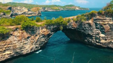 Berrak mavi gökyüzü altında yemyeşil ve engebeli uçurumlarla çevrili turkuaz suların üzerinde doğal bir kaya kemeri barındıran çarpıcı bir kıyı manzarası..