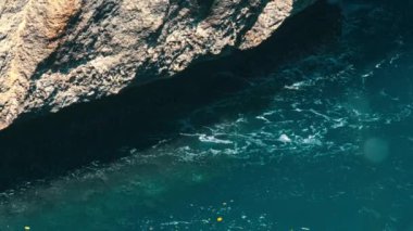 Açık mavi suyu olan kayalık bir sahil şeridine yakın çekim. Güneş ışığı suyun yüzeyinden yansıyarak parıldayan bir etki yaratır. Kayalar pürüzsüz suyla çelişen dokulu ve engebelidir..