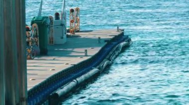 Turkuaz bir denize uzanan ahşap bir rıhtım, yeşil bir çöp kutusu ve görünür güvenlik ekipmanıyla. Su durgun ve mavi gökyüzünü yansıtıyor..