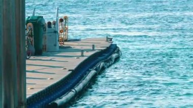 Berrak mavi suya uzanan ahşap bir rıhtım manzarası, yeşil bir çöp kovası ve rıhtımda görülebilen bazı ekipmanlar..