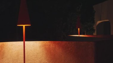 Modern açık hava lambalarının kıvrımlı bir duvara sıcak ışık saçtığı huzurlu bir gece sahnesi. Lambaların, uzayın atmosferini arttıran eşsiz bir konik tasarımı var..