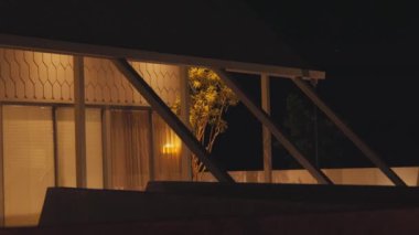 Geceleri sıcak ışıklarla aydınlatılmış modern bir ev. Yapıda büyük pencereler ve dışarıda görünen bir ağaç ile benzersiz bir mimari tasarım yer alıyor. Sahne rahat ve davetkar bir atmosfer sunuyor..