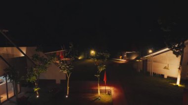 Yumuşak ışıklarla aydınlatılmış çok sayıda binanın yer aldığı modern mimari manzaranın huzurlu bir gece manzarası. Yemyeşil çimenler ağaçlar ve dekoratif aydınlatmalarla kaplı. Huzurlu bir ortam yaratıyor..