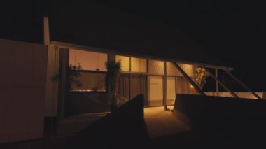 Geceleri aydınlatılan, geniş pencereler ve gösterişli bir tasarım sergileyen modern bir ev. Dış yüzeyde, girişe giden karanlık bir yolu olan, yeşilliklerle çevrili minimalist bir estetik var..
