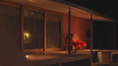 Geceleri modern bir dış mekan, büyük cam kapılar ve iç ışıklardan gelen sıcak bir parıltı. İki parlak kırmızı sandalye verandada, yeşilliklerle çevrili..