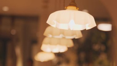 Beyaz gölgeli, zarif kolye ışıkları serisi. Kapalı bir mekanı hafifçe aydınlatıyor. Işıklar arka arkaya dizilmiş, sıcak bir ortam yaratıyor..