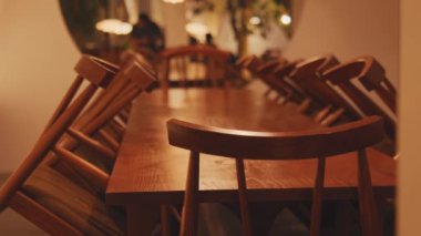 Üzerinde sandalyeler olan ahşap bir yemek masası, rahat ve hafif aydınlatılmış bir restoranda kurulmuş. Ormanın sıcak tonları, arka planda bulanık figürlerle davetkar bir atmosfer yaratıyor..