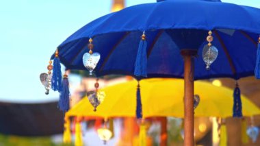 Parlak mavi gökyüzüne karşı dizilmiş püsküllü ve süslemeli renkli şemsiyeler. Ön planda mavi bir şemsiye yer alırken, arka planda sarı bir şemsiye bulanıklaşarak canlı ve şenlikli bir atmosfer yaratıyor..
