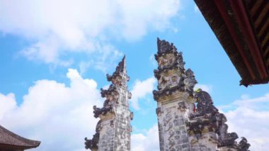 Karmaşık oymaları olan iki süslü taş kule, kabarık bulutlu parlak mavi gökyüzüne karşı kurulmuş. Kuleler geleneksel Bali tapınağı mimarisinin bir parçası. Kültür mirası sergileniyor..