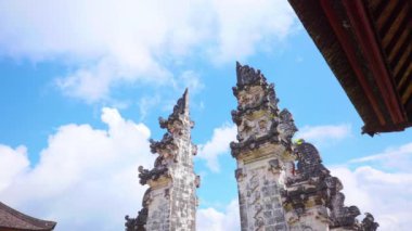 Karmaşık oymaları olan iki antik taş kule, parlak mavi gökyüzüne karşı kabarık beyaz bulutlarla. Kuleler, bölgenin eşsiz mimari tarzını sergileyen geleneksel Bali tapınağının bir parçası..