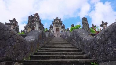 Geleneksel bir Bali tapınağına çıkan heybetli bir taş merdiven. Mavi gökyüzünün altında kabarık bulutlarla dolu karmaşık oymalar ve yemyeşil bir yapı..