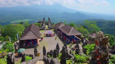 Tepedeki tapınak kompleksinin panoramik görüntüsü, yemyeşil ve arka planda dağlarla çevrili. Geleneksel Bali mimarisi ve taş oymalarıyla bölgeyi gezen ziyaretçiler görülüyor.
