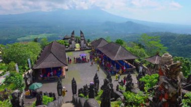 Endonezya, Bali 'de yemyeşil ve dağlarla çevrili bir tapınak kompleksinin panoramik görüntüsü. Ziyaretçiler, ön planda geleneksel mimari ve heykellerin yer aldığı bir kültür etkinliğine katılıyorlar..