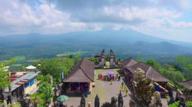Dağdaki yemyeşil bir tapınak kompleksinin panoramik görüntüsü ve bulutlu bir gökyüzünün altında uzak bir volkan. İnsanlar tapınak bölgesinde toplandı, kültürel aktiviteler sergiliyorlar..